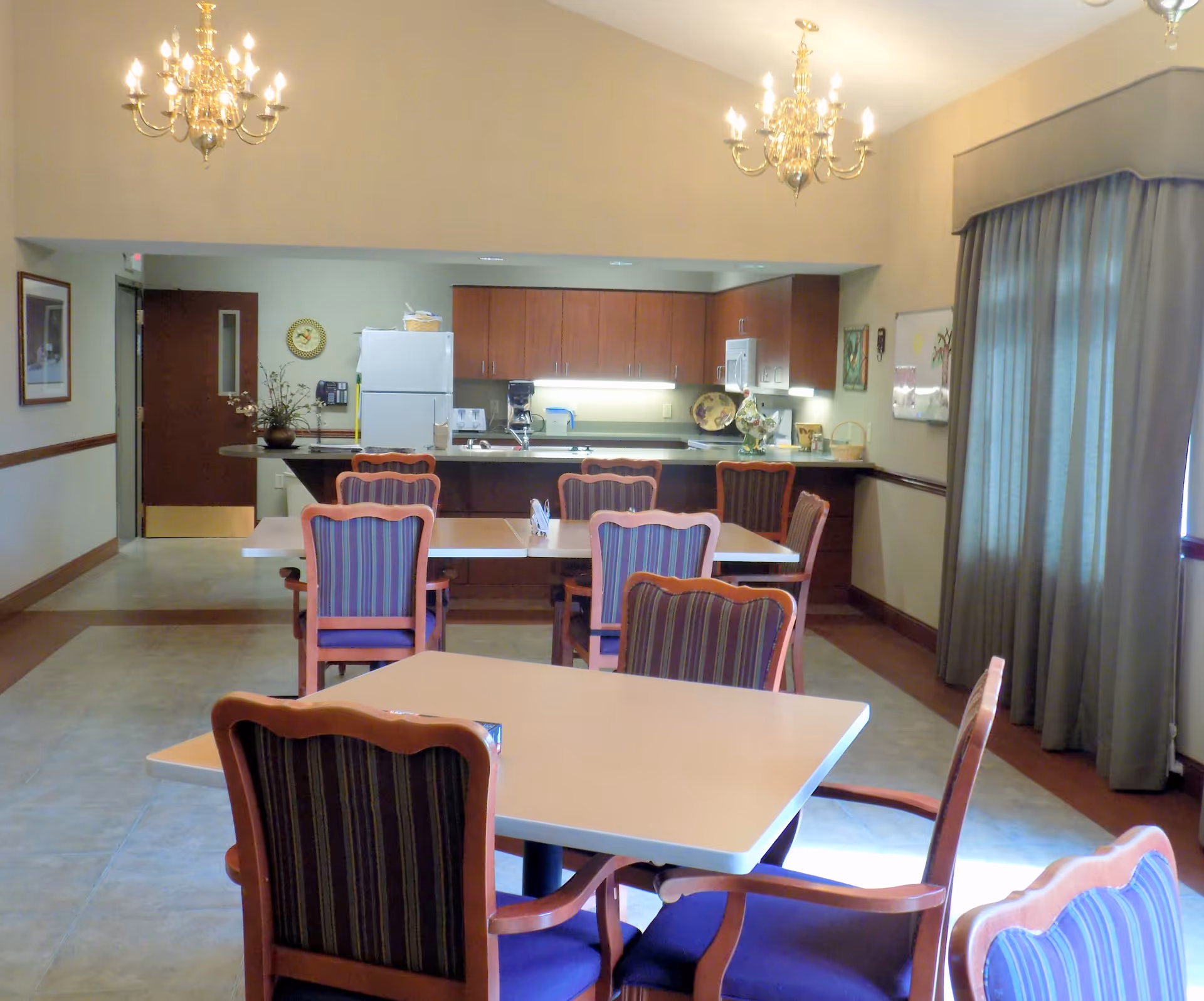 A dining area with several tables and chairs featuring purple striped upholstery. In the background, there is a kitchen area with wooden cabinets, a white refrigerator, a microwave, and a coffee maker. Two chandeliers hang from the ceiling, and a large window with closed curtains is on the right side.