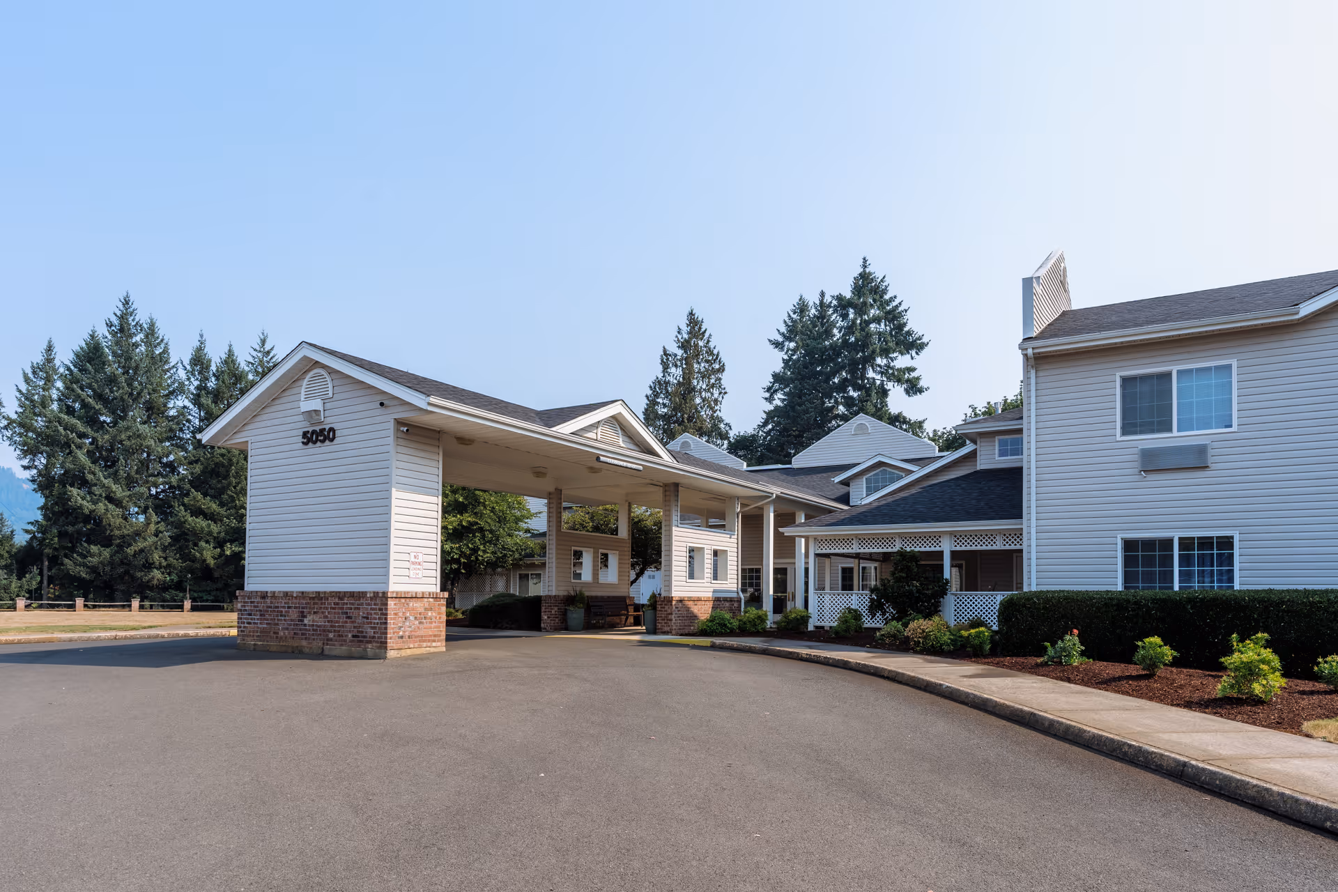 Exterior view of Wiley Creek Senior Living facility showing a covered entrance with the number 5050 on the side, surrounded by trees and landscaping under a clear sky.