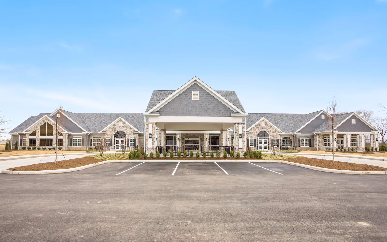 Front exterior of a single-story senior living building with a covered entrance, stone and siding facade, and empty parking spaces.