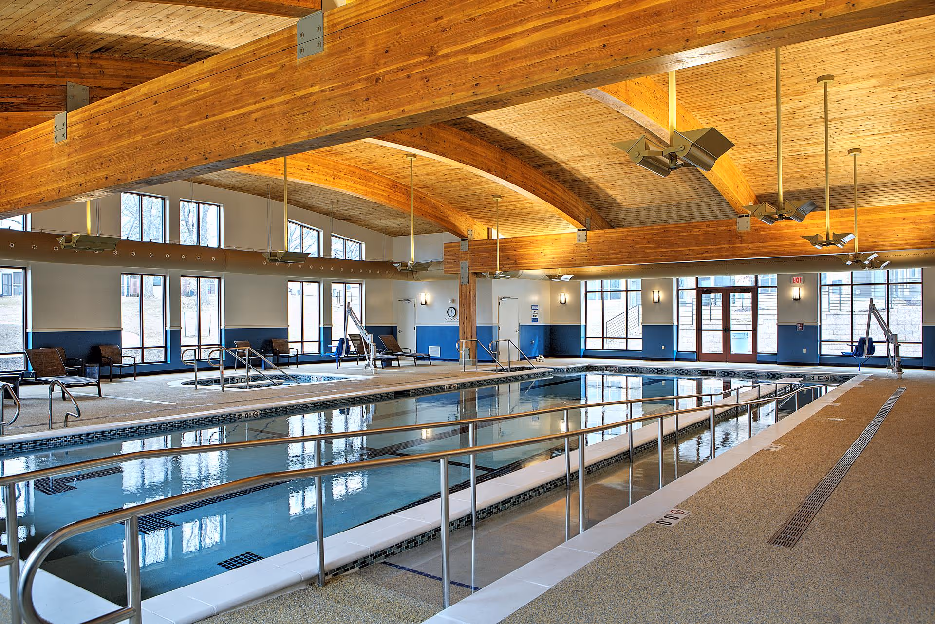 Indoor swimming pool area with arched wooden ceiling, handrails, lounge chairs and large windows.