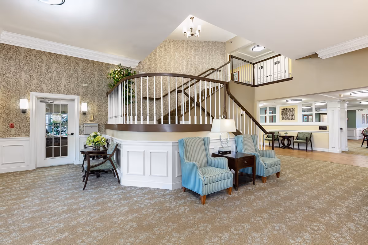A spacious and well-lit interior common area featuring a carpeted floor, two blue upholstered armchairs with a small wooden table and lamp between them, a curved staircase with white spindles and dark handrails leading to an upper floor, patterned wallpaper, and additional seating and decorative elements in the background.