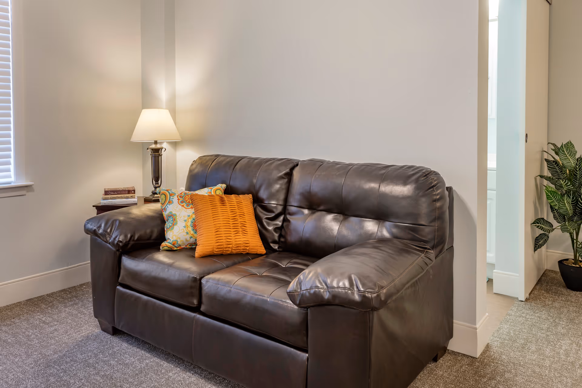 A cozy living room area featuring a dark brown leather loveseat with two decorative pillows, one orange and one patterned. A side table with a lamp and a few books is placed next to the loveseat. There is a window with blinds on the left and a potted plant near an open doorway on the right.