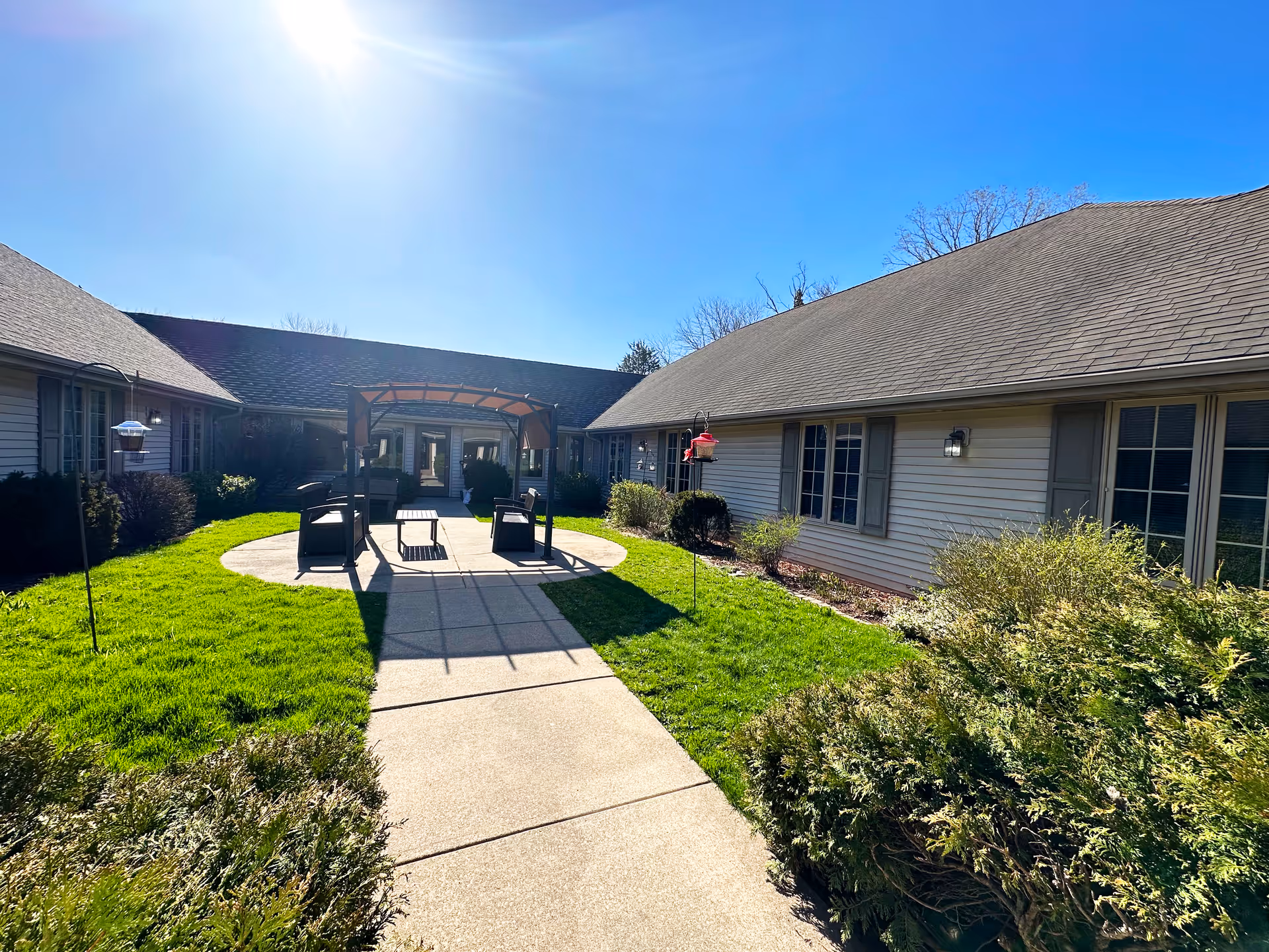 Outdoor courtyard area at Wyndemere Memory Care featuring a paved walkway leading to a shaded seating area with chairs and a table. The courtyard is surrounded by single-story buildings with windows and greenery, under a clear blue sky with bright sunlight.