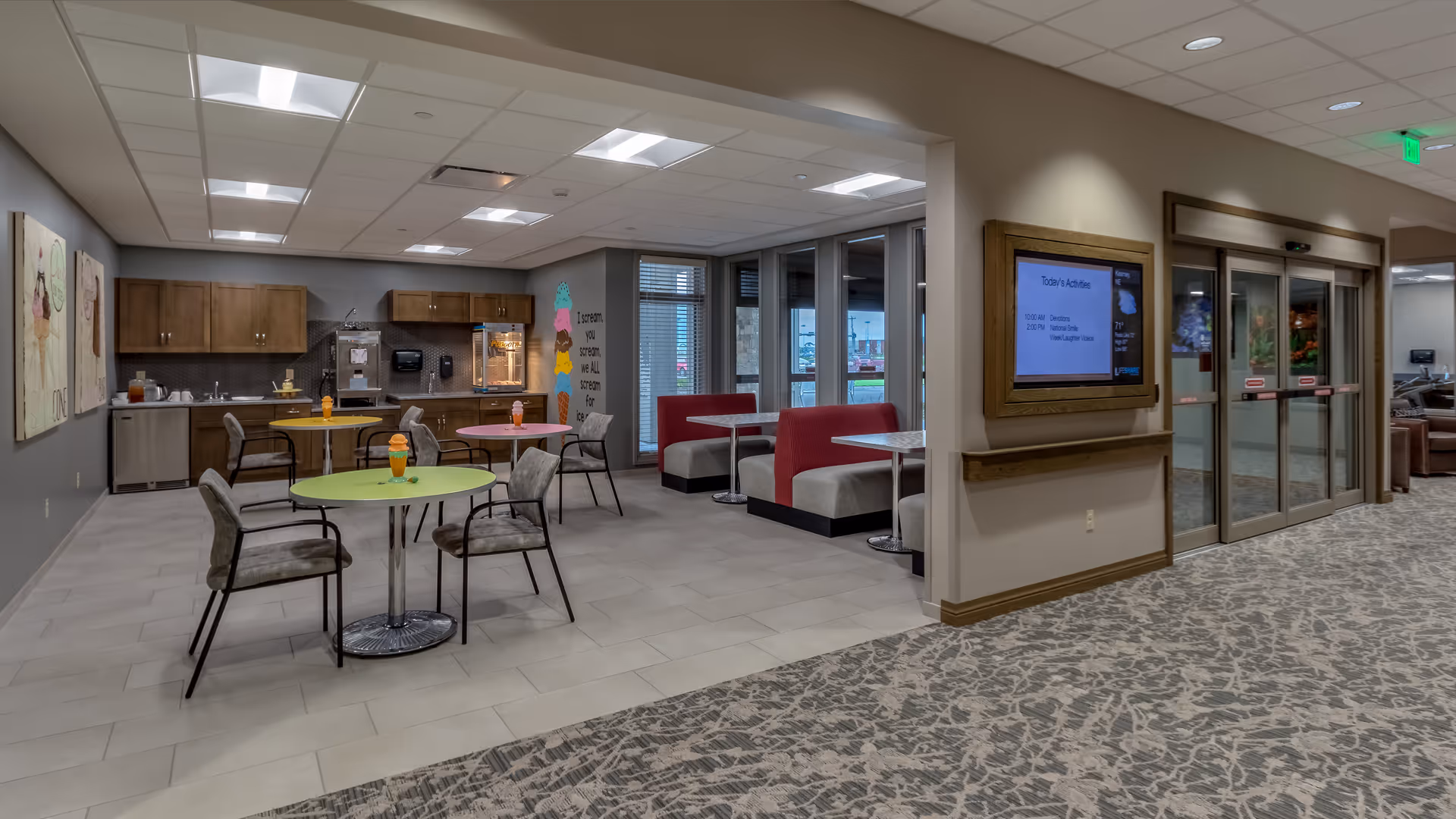 Interior view of a senior living facility common area with small round tables and chairs, a kitchenette with cabinets and a popcorn machine, red and gray booth seating near large windows, and an entrance with automatic sliding glass doors next to a digital bulletin board displaying today's activities.