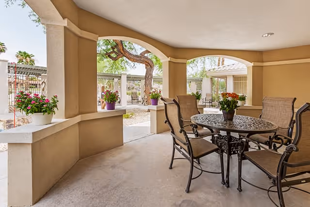 Covered outdoor patio with a round metal table, four chairs, and potted flowers under arched openings overlooking a courtyard.