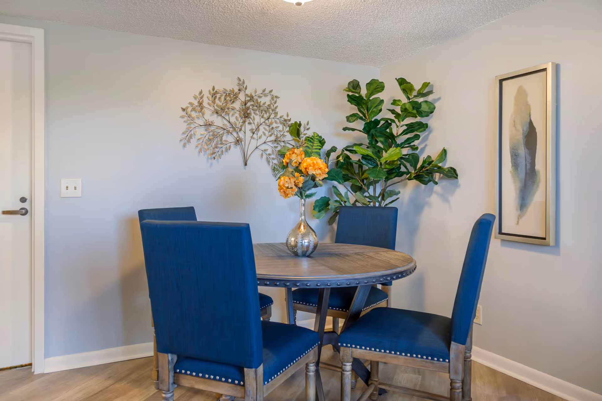 A small dining area with a round wooden table surrounded by four blue upholstered chairs. On the table is a silver vase with orange flowers. The walls are light gray with a metal wall decoration shaped like leaves and a framed abstract artwork. There is a large green leafy plant in the corner and a white door to the left.