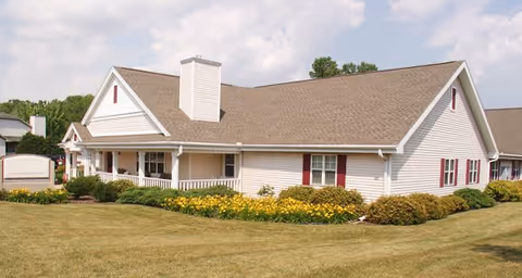 Exterior view of a single-story assisted living facility building with beige siding, a brown shingled roof, white trim, and red shutters. The building is surrounded by a well-maintained lawn, yellow flowers, and shrubs under a partly cloudy sky.