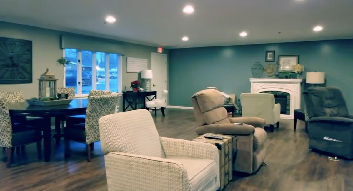 A cozy living room area with several upholstered armchairs and recliners arranged around a white decorative fireplace. There is a wooden dining table with patterned chairs near a window, and the room has wood flooring and soft lighting from ceiling fixtures and lamps.