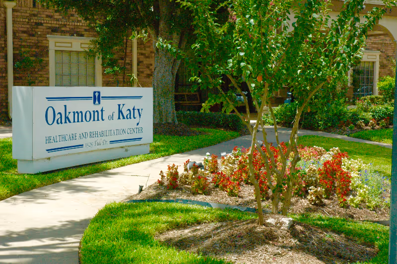 Landscaped entrance with a white sign reading "Oakmont of Katy Healthcare and Rehabilitation Center" in front of a brick building.