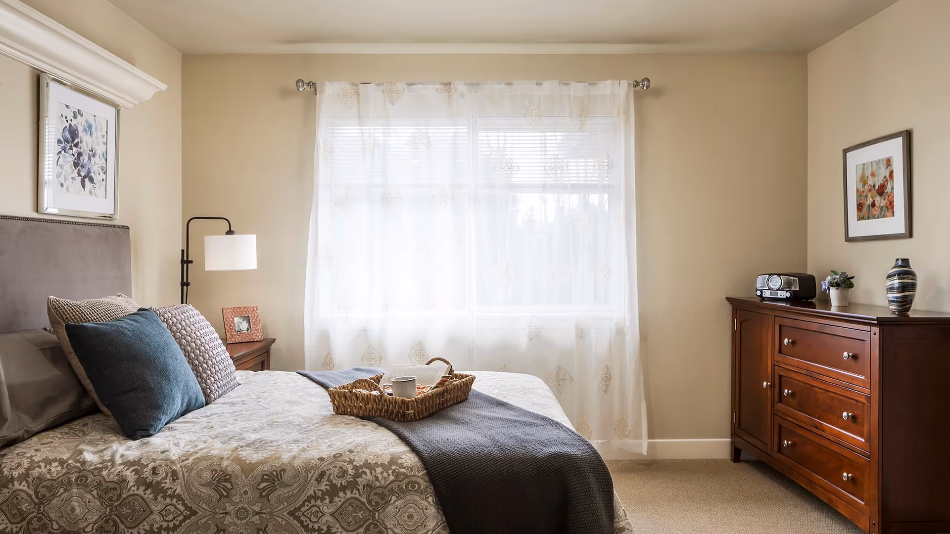 A cozy bedroom with a neatly made bed featuring patterned bedding and multiple pillows. A wicker tray with a cup and a small item sits on the bed. To the left of the bed is a nightstand with a lamp and a framed photo. A large window with sheer curtains allows natural light to fill the room. On the right side, there is a wooden dresser with a radio, a small plant, and a decorative vase. Two framed floral artworks hang on the walls.