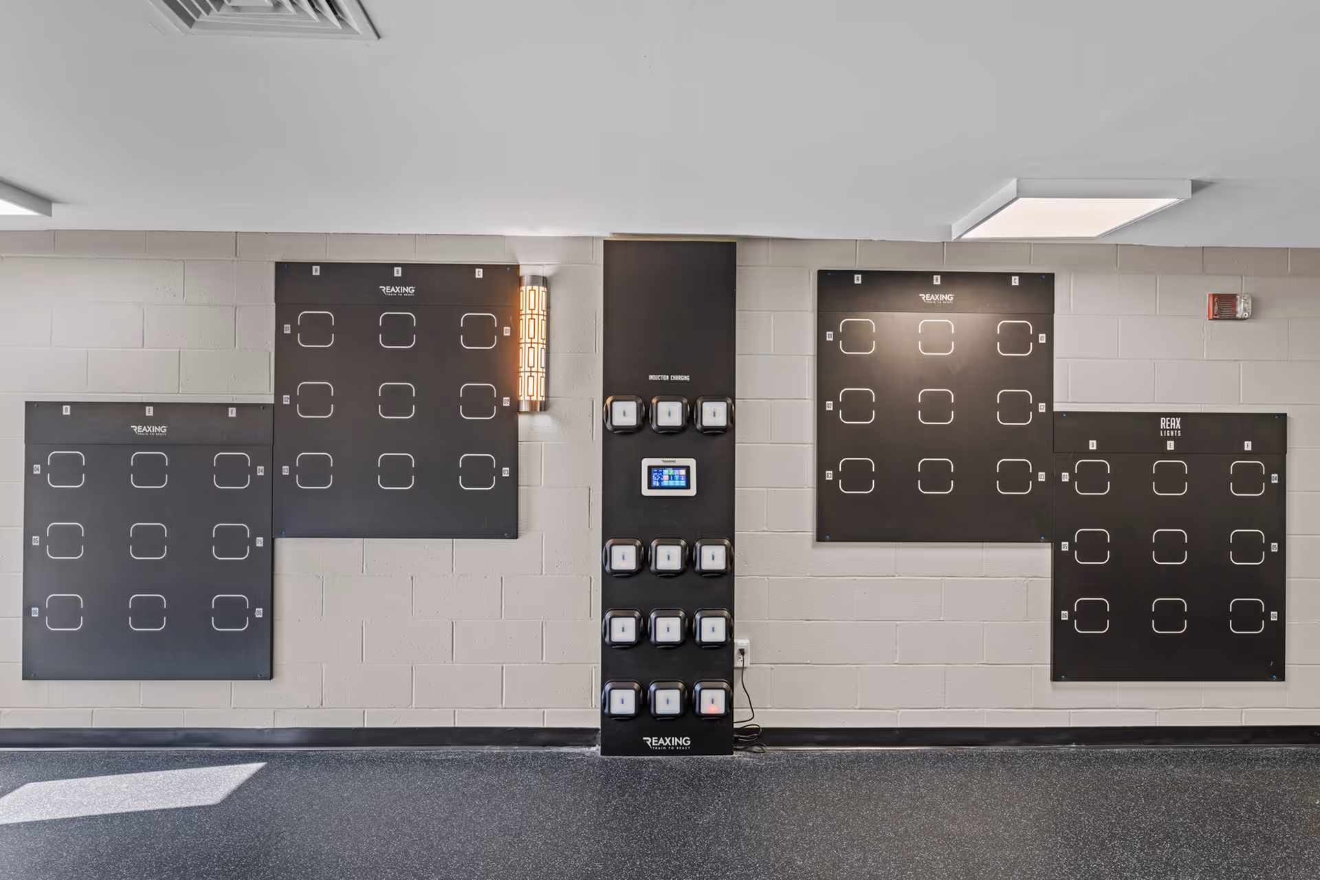 A wall-mounted exercise setup with multiple black panels featuring square outlines for hand or foot placement, and a central column with various mounted devices and a small touchscreen. The room has light gray painted cinder block walls and a dark speckled floor.