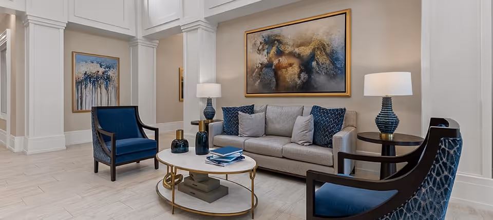 A stylish living room area with a beige sofa adorned with blue and gray pillows, two blue upholstered armchairs with dark wooden frames, a white oval coffee table with decorative items and books, two matching blue table lamps on round side tables, and abstract artwork on the walls.