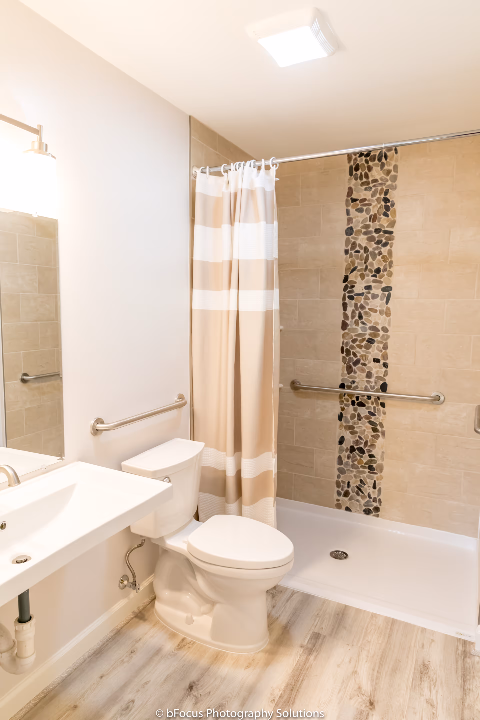 A clean and modern bathroom featuring a white toilet, a white sink with a silver faucet, a beige and white striped shower curtain, and a walk-in shower with beige tiles and a vertical strip of pebble-style mosaic tiles. The bathroom has light wood flooring and grab bars installed near the toilet and inside the shower for accessibility.