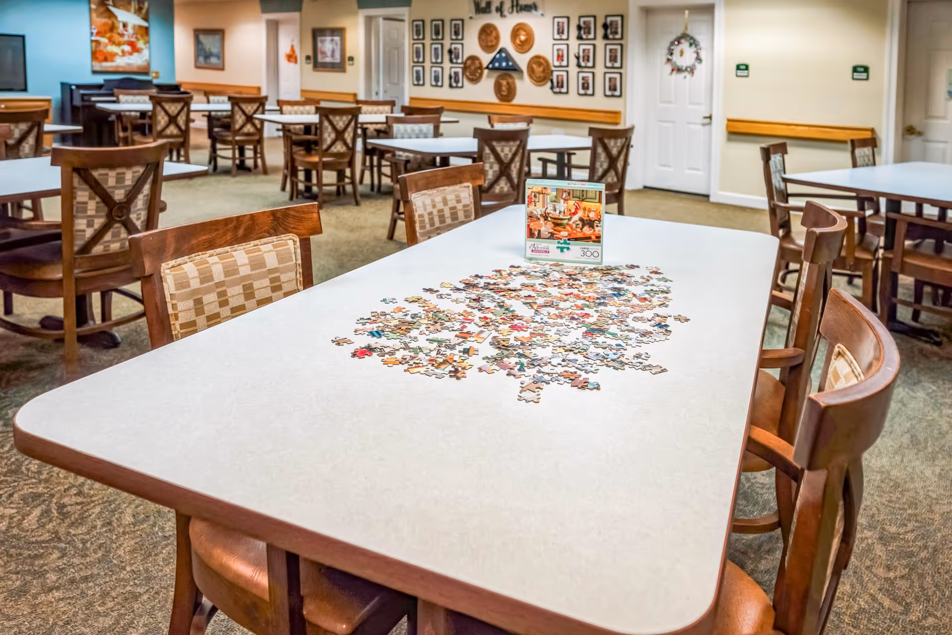A senior living facility common room with multiple tables and chairs. One table in the foreground has an incomplete jigsaw puzzle spread out with the puzzle box standing upright. The room has carpeted floors, wall decorations including framed pictures and plaques, and a piano in the background.