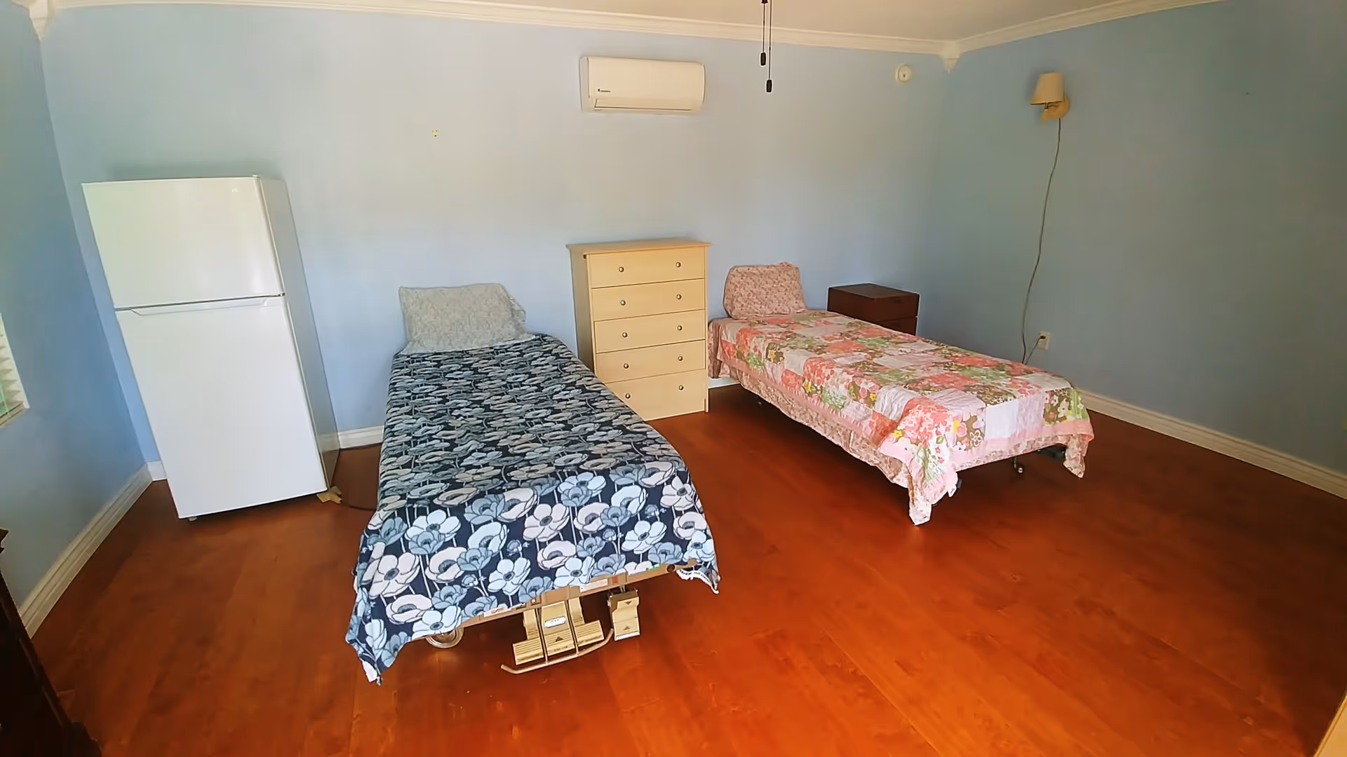 A simple bedroom with two single beds, each covered with different floral bedspreads. Between the beds is a small chest of drawers. To the left of the beds is a white refrigerator. The room has light blue walls, wooden flooring, a wall-mounted air conditioner, and a wall lamp with a visible cord.