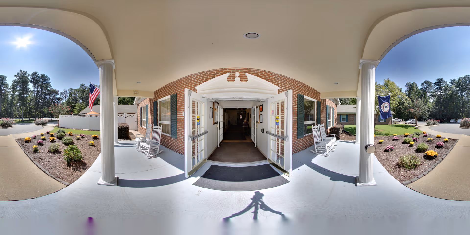 Entrance to Dominion Village at Williamsburg showing a covered porch with white columns and rocking chairs on either side of double glass doors. There are landscaped flower beds with colorful flowers and an American flag on the left side and another flag on the right side. Trees and a parking area are visible in the background under a clear blue sky.