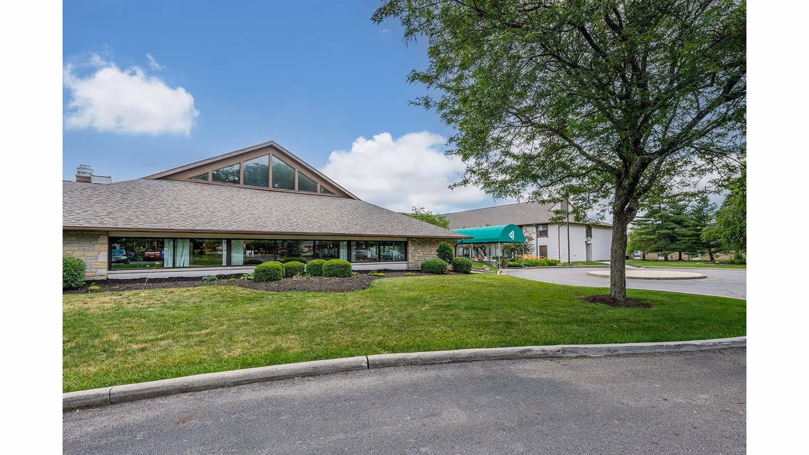 Exterior view of The Village of Westerville facility showing a single-story building with large windows, a green awning over the entrance, a well-maintained lawn, bushes, a tree, and a clear blue sky with some clouds.