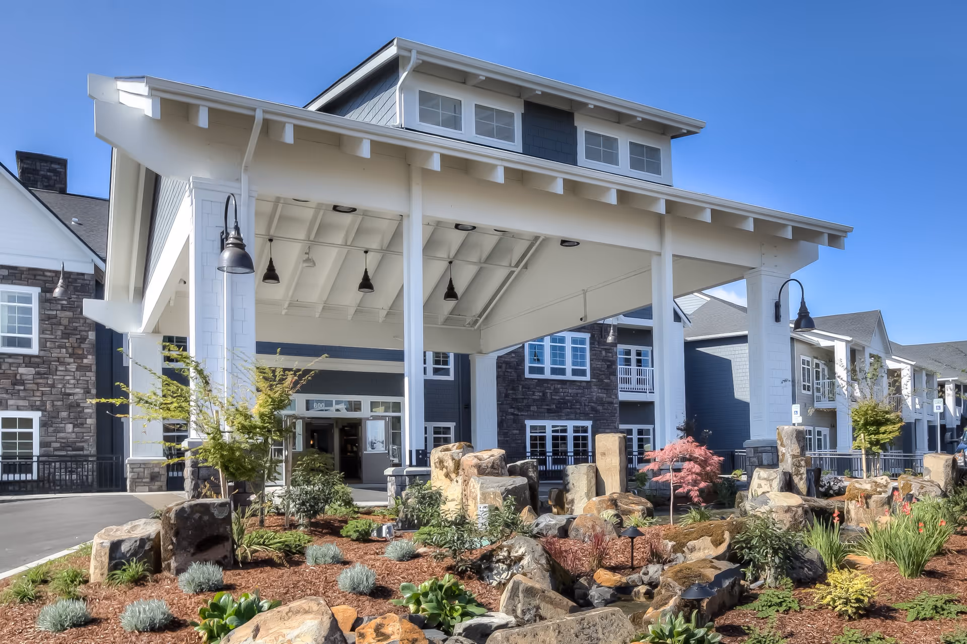 Entrance of a senior living facility named Waterford Grand featuring a large covered driveway with white pillars, landscaped garden with rocks and plants in the foreground, and multi-story buildings with stone and siding exteriors in the background under a clear blue sky.