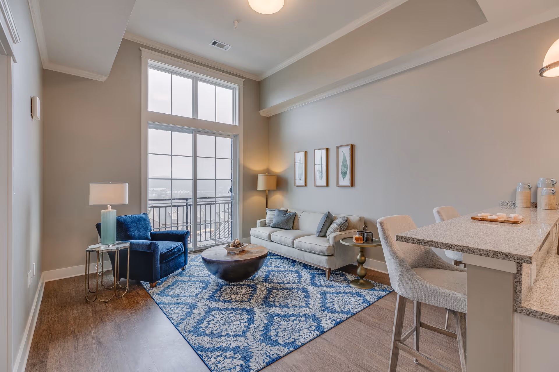 A modern living room with a large window and glass door leading to a balcony. The room features a blue patterned rug, a beige sofa with cushions, a round wooden coffee table, a blue armchair with a side table and lamp, and a kitchen counter with two upholstered bar stools. Three framed botanical prints hang on the wall above the sofa.