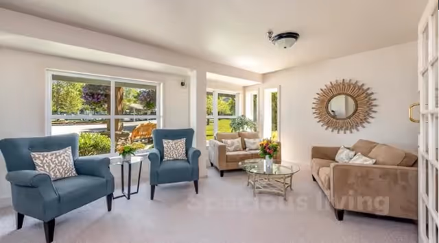 Bright living room with two blue armchairs, beige sofas, a glass coffee table and large windows overlooking greenery.
