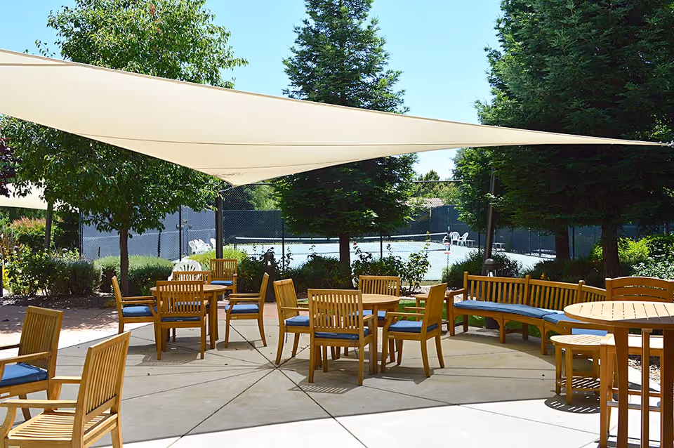 Outdoor seating area with wooden chairs and tables arranged on a concrete patio under large white shade sails. Surrounding the patio are green trees and bushes, with a tennis court visible in the background behind a chain-link fence.