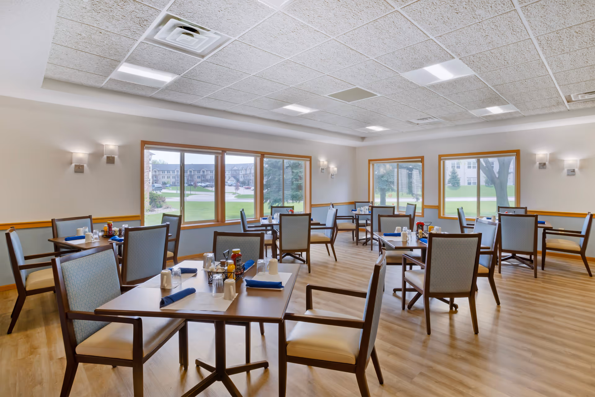 A bright dining room with multiple square tables set with blue napkins, white mugs, glasses, and condiment holders. The room has large windows with wooden frames letting in natural light and providing a view of green lawns and buildings outside. The floor is wooden, and the walls are painted in light colors with wall-mounted lights.