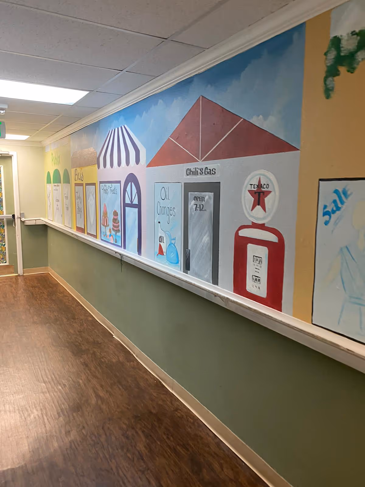 Interior hallway with a colorful mural of storefronts painted along the wall and a wood-look floor.