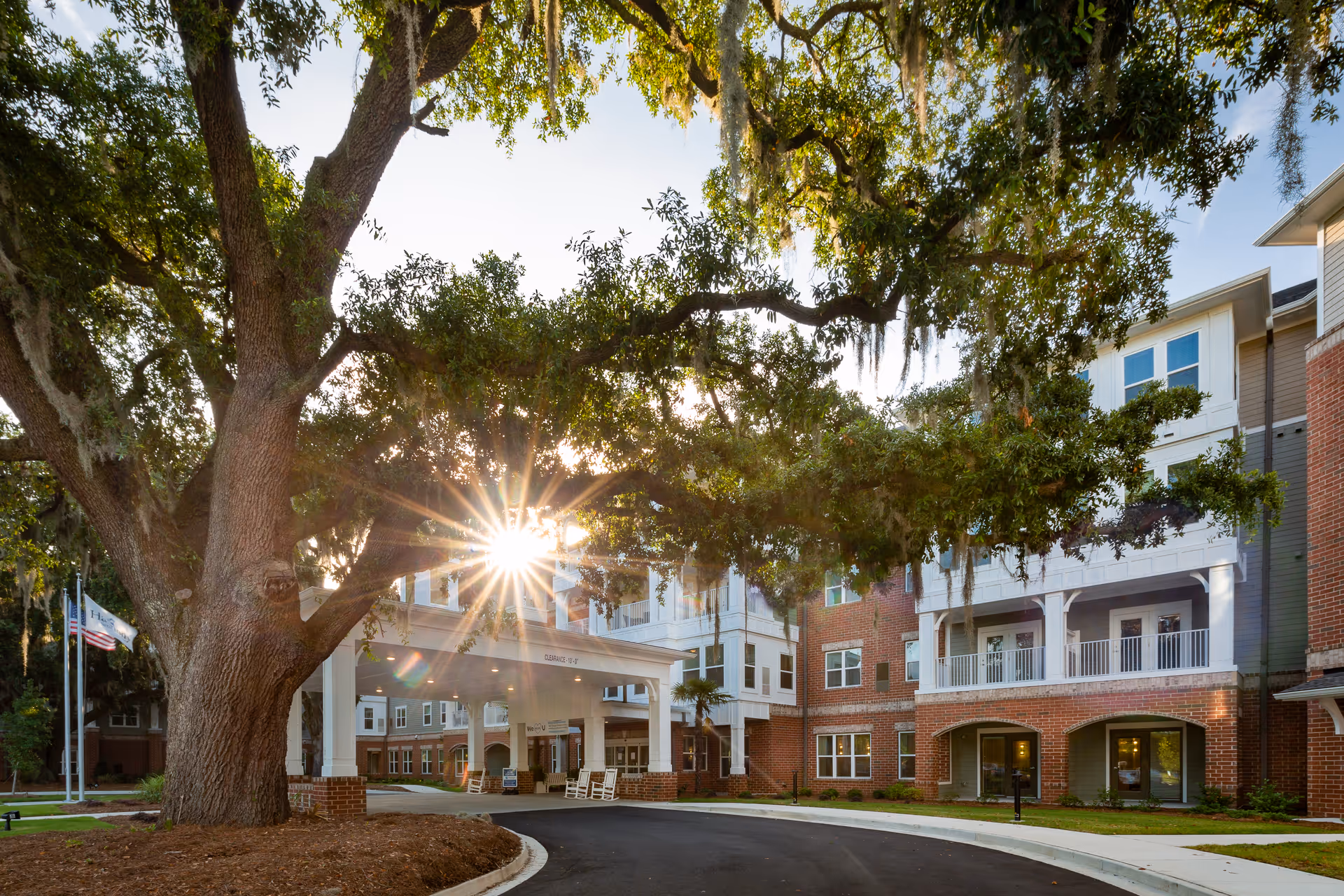 Front entrance of a senior living building with a large oak tree and sunlight shining through its branches.