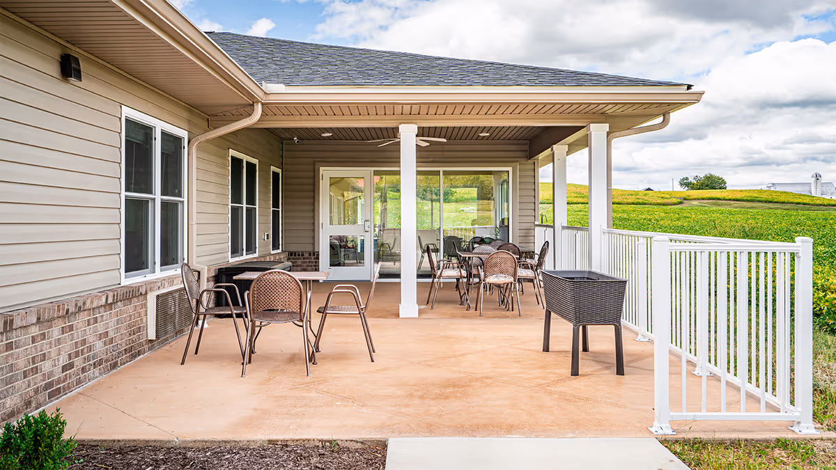 Covered outdoor patio with tables and chairs attached to a light-colored building overlooking grassy fields.