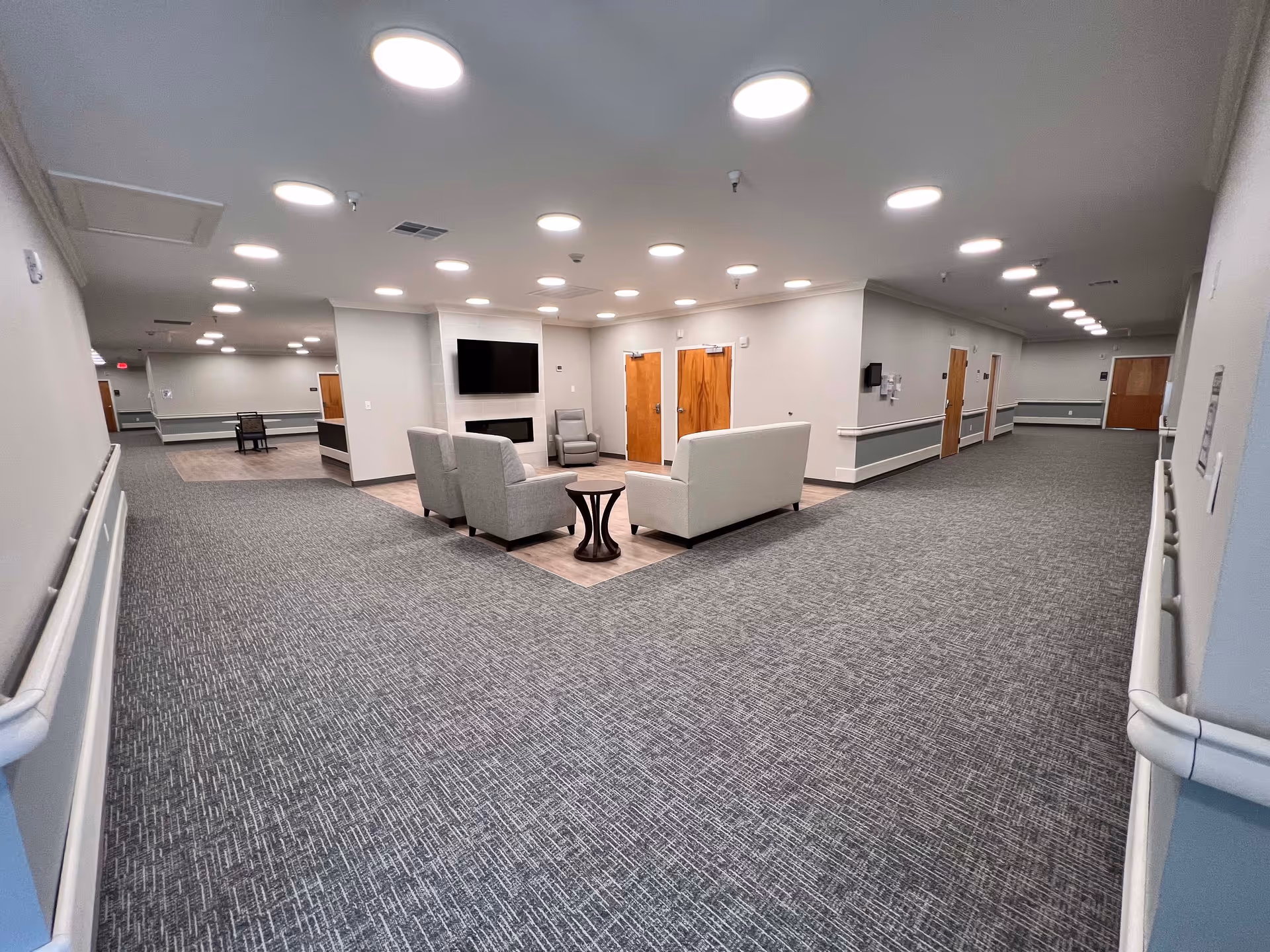 Spacious, brightly lit senior living hallway with a seating area and TV in the middle and multiple doors along the corridor.