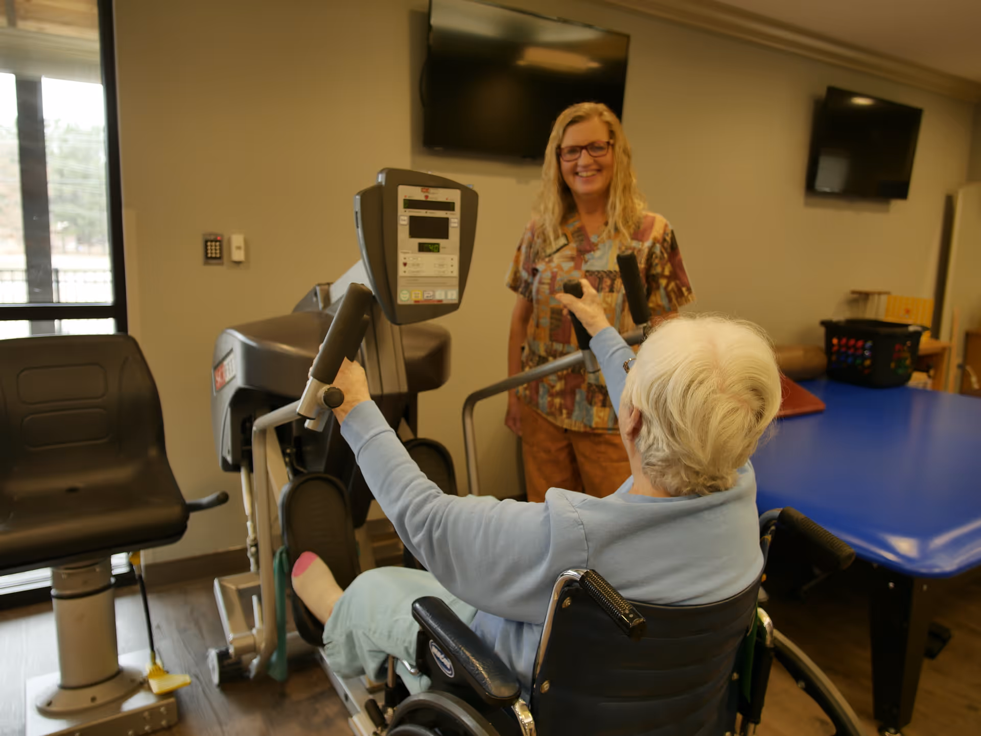 An elderly person in a wheelchair using an exercise machine in a rehabilitation room, assisted by a smiling staff member wearing scrubs. The room has exercise equipment, a blue therapy table, and two wall-mounted televisions.