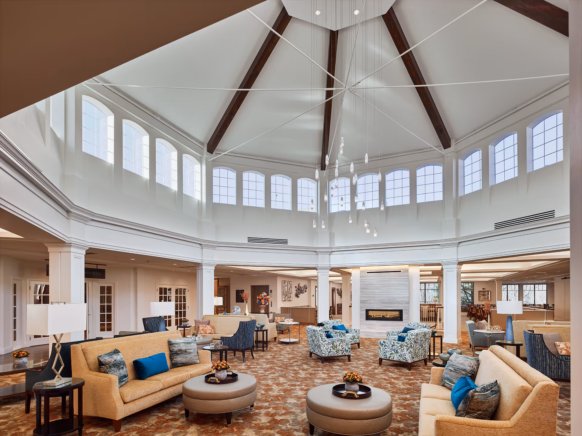 Spacious and well-lit senior living facility common area with high vaulted ceiling featuring wooden beams and multiple large windows. The room is furnished with beige sofas, patterned armchairs, round ottomans, side tables with lamps, and a modern fireplace in the center. The carpet has a warm, patterned design and the overall atmosphere is inviting and comfortable.