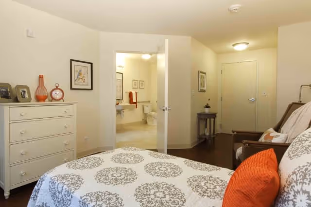 A bedroom with a bed featuring a white and gray patterned bedspread and orange pillows. To the left is a white dresser with decorative items on top. The room has light-colored walls and wooden flooring. An open door reveals a bathroom with a toilet and sink visible. A small table with a plant is near the closed door in the background.