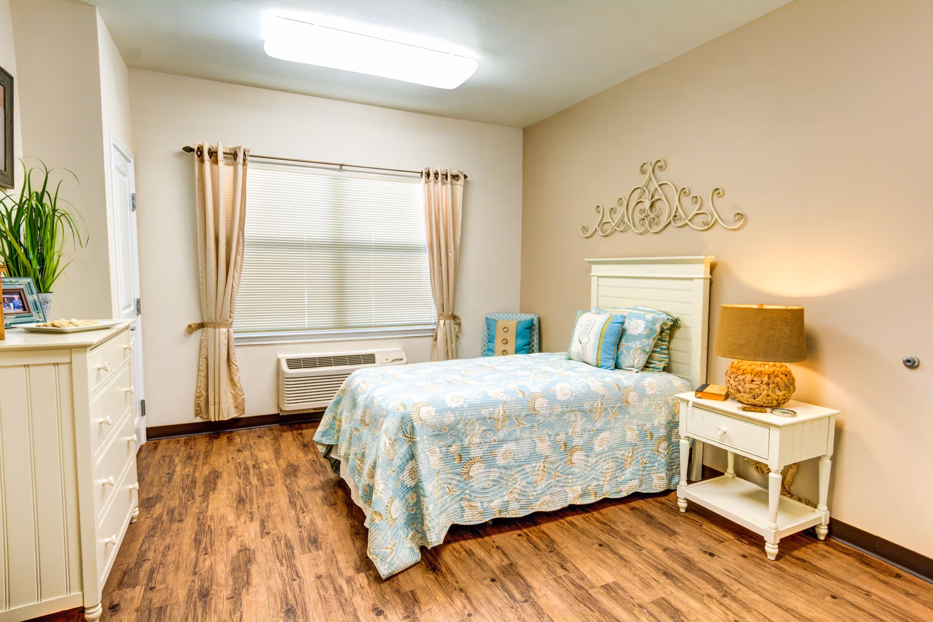 A cozy bedroom with a single bed covered in a blue patterned quilt and matching pillows. There is a white nightstand with a lamp and a small book next to the bed. A decorative wall piece hangs above the headboard. The room has wood flooring, beige walls, a window with beige curtains, and a white dresser with a plant and framed photos on top.