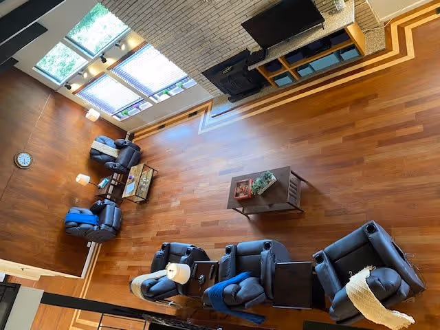A spacious living room with wooden flooring and a high ceiling. The room features several dark blue recliner chairs arranged around a central wooden coffee table with a plant and decorative items. There is a fireplace with a TV mounted above it on one wall, and large windows with blinds letting in natural light. A clock is mounted on a wooden wall, and there are small lamps on side tables next to the chairs.