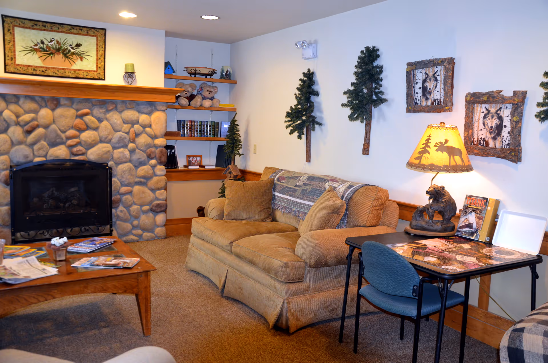 Cozy living room with a stone fireplace, cushioned sofa, coffee table, wall-mounted tree decorations, and a small side table with a lamp.