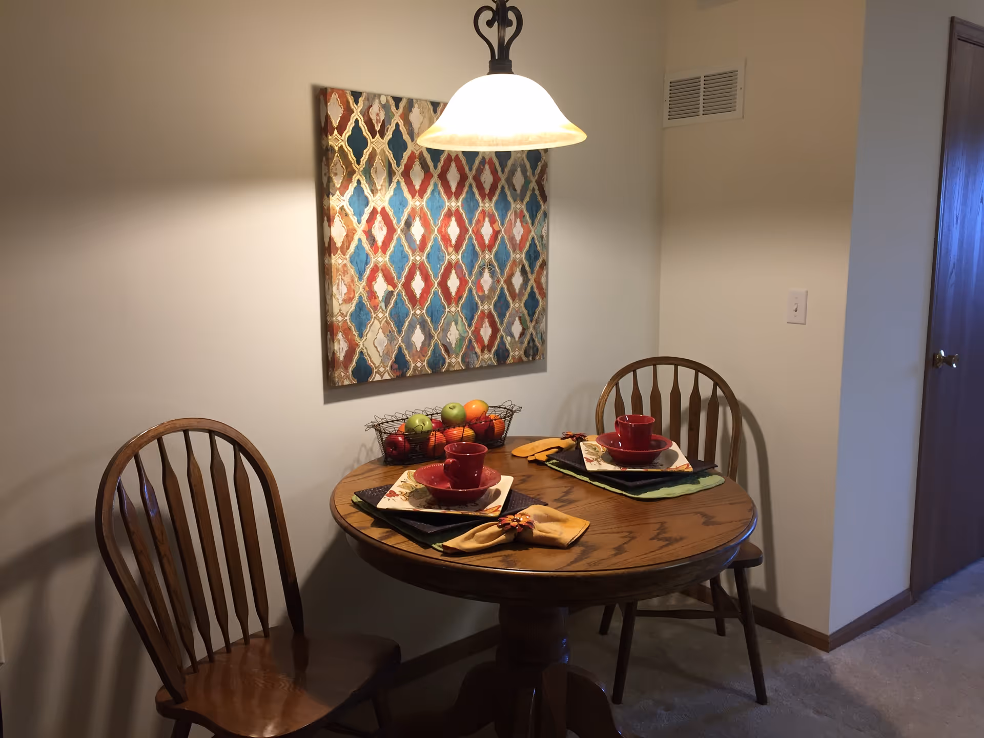 Small dining nook with a round wooden table set for two, two wooden chairs, a hanging light, and colorful wall art on the wall.