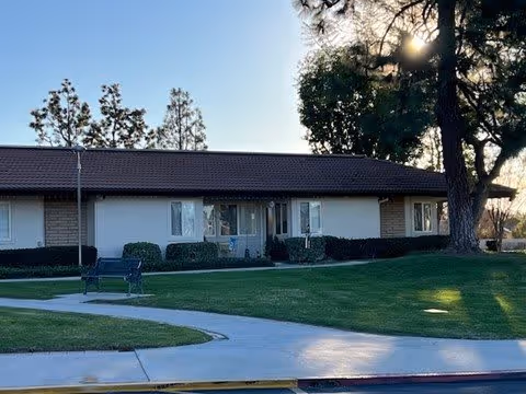 Single-story residential building with a manicured lawn, walkway, bench, and large trees in front at sunset.
