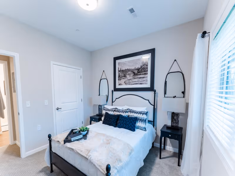 A cozy bedroom with a black metal bed frame, white bedding, and decorative pillows. There are two black nightstands on either side of the bed, each with a lamp and a hanging oval mirror above. A framed black and white picture hangs above the bed. A window with white blinds and curtains is on the right side, and a door is visible on the left side of the room.
