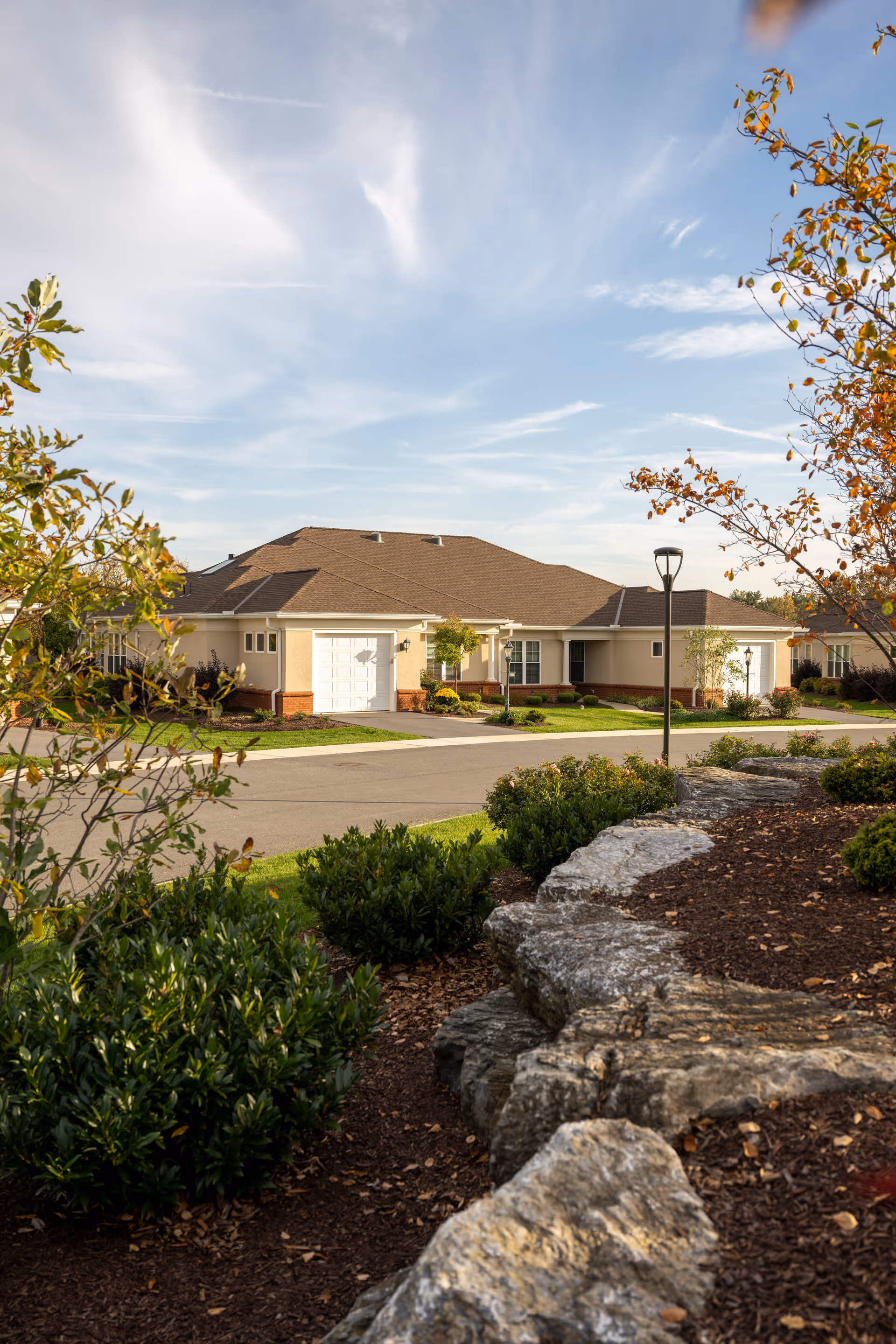 Single-story senior living building with garages, landscaped grounds and a rock wall in the foreground under a clear blue sky.