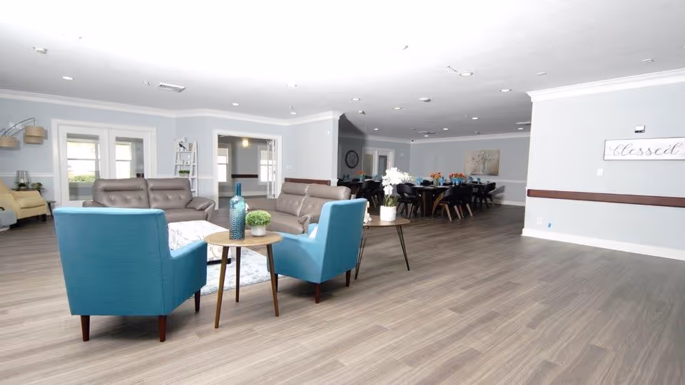 A spacious, well-lit common area in an assisted living facility featuring two blue armchairs and two brown sofas arranged around a small round wooden table with decorative vases and plants. In the background, there is a dining area with multiple tables and chairs, light gray walls, and wood flooring. A wall sign reads 'Blessed'.