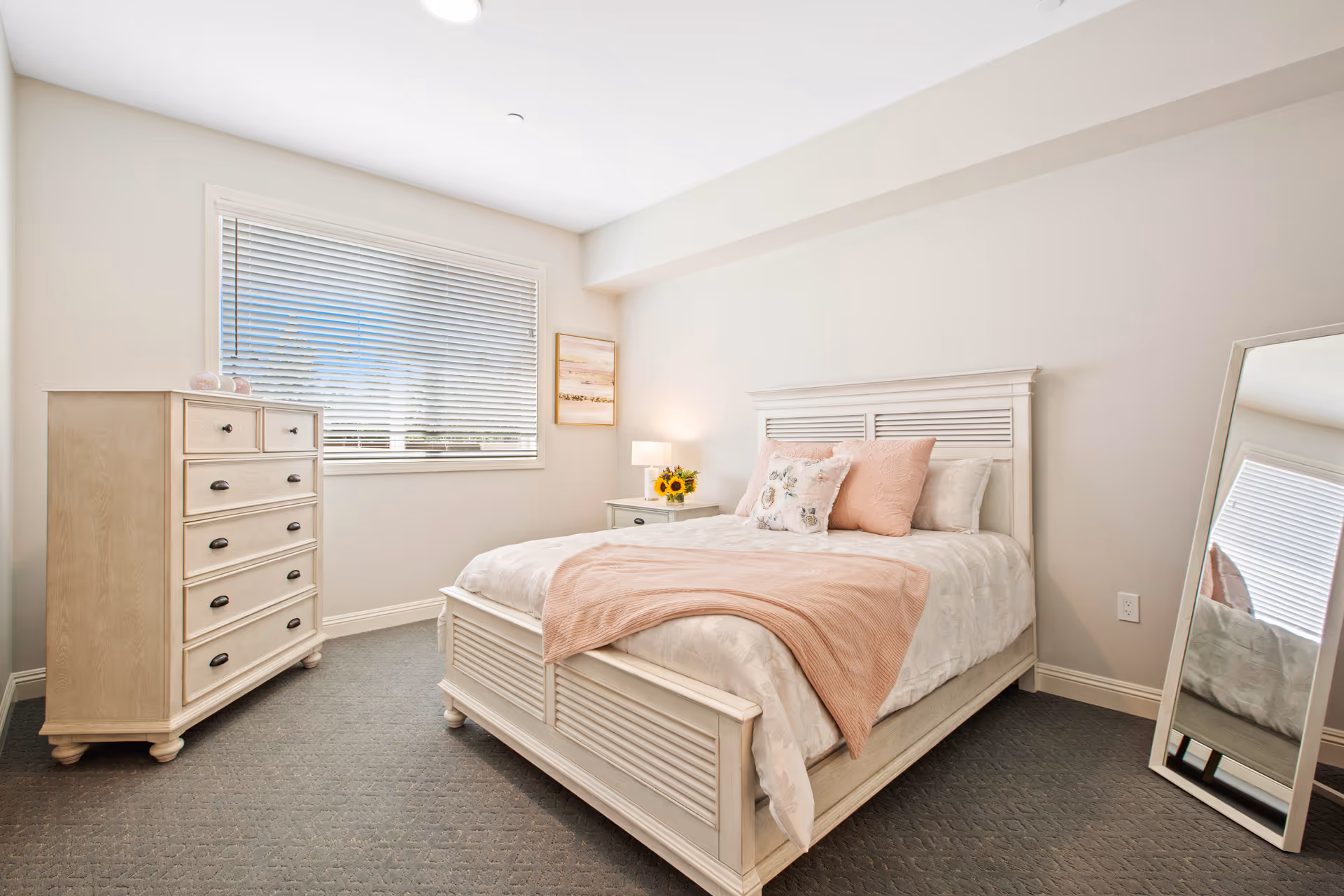 Sunlit bedroom with a white wooden bed dressed in pale pink and white linens, a matching dresser, nightstand, and a leaning full-length mirror.