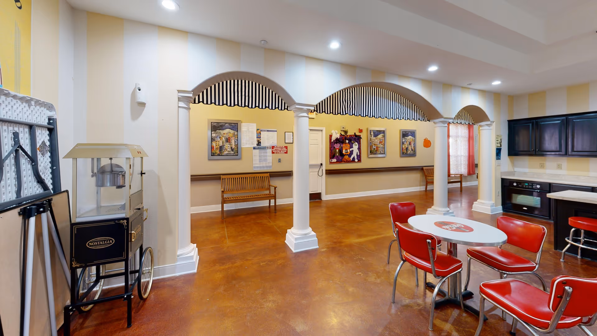 Interior view of a senior living facility common area with a retro-style popcorn machine on the left, three white columns with striped awnings, a small round table with four red chairs, and a kitchenette with dark cabinets and an oven. The walls have light yellow and white vertical stripes, and there are benches and framed pictures in the background.