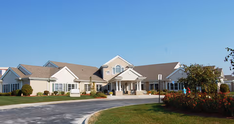 Front exterior view of a single-story senior living facility building with a covered entrance, surrounded by a curved driveway, green lawn, and some bushes and trees under a clear blue sky.