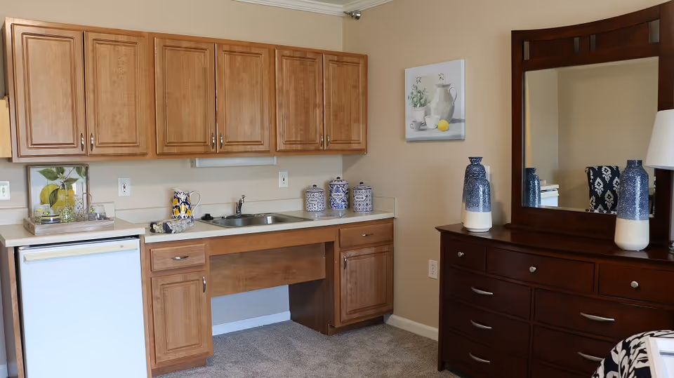 Studio-style interior with a kitchenette (cabinets, sink, mini-fridge) and a dark wooden dresser with mirror and decorative vases.