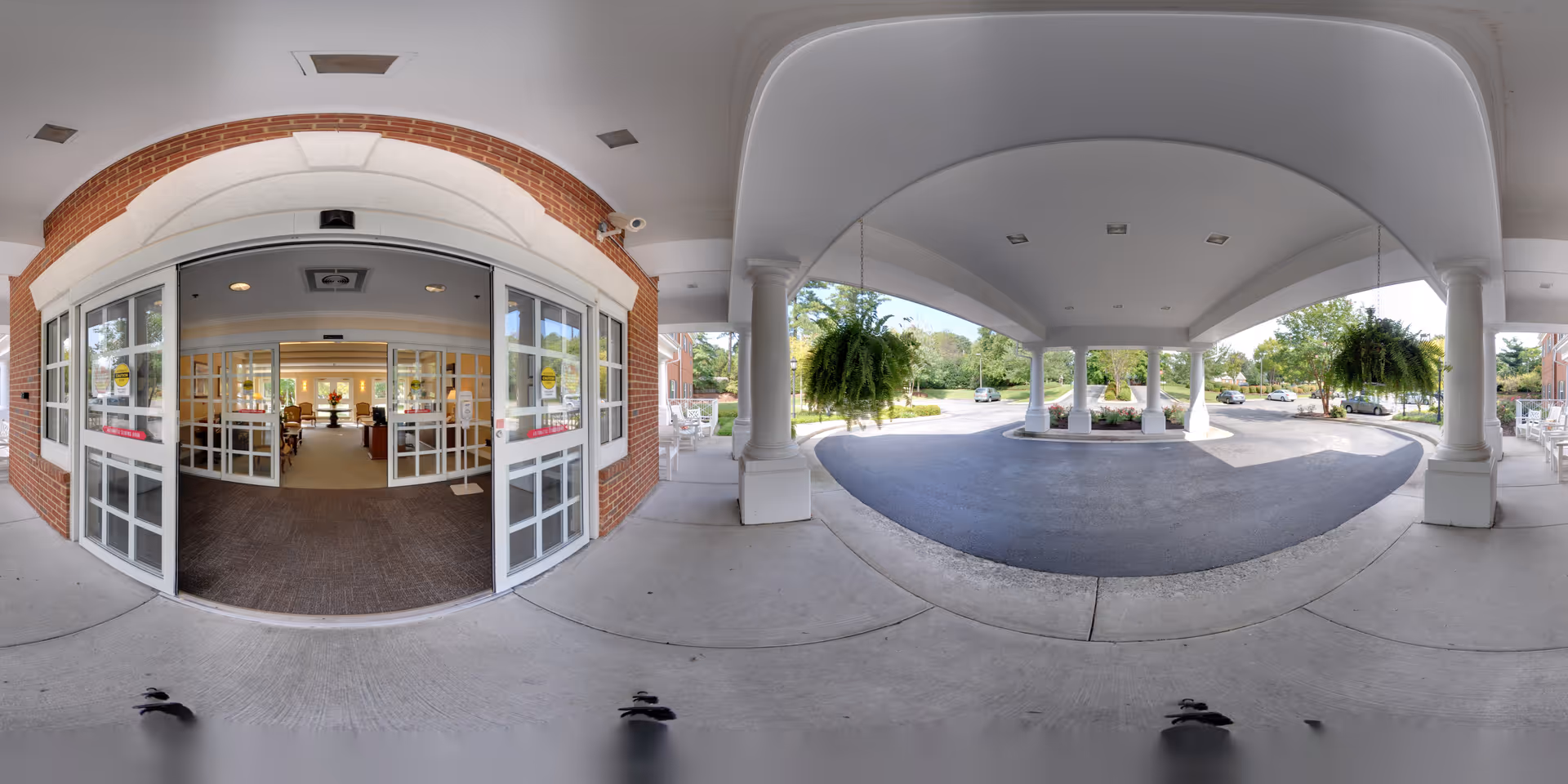 Covered entrance area of a senior living facility with white pillars and hanging plants. The entrance has large glass double doors leading into a well-lit interior space with seating visible inside. Outside, there is a driveway for vehicles to pull up under the covered area, with trees and parked cars in the background.