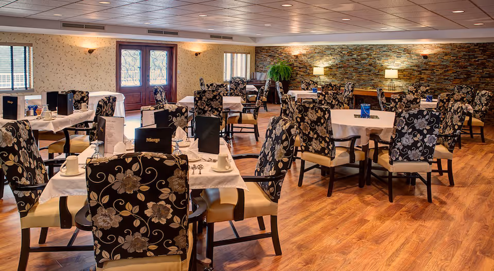 Spacious dining room with round tables set for meals and patterned upholstered chairs on wood floors and a stone accent wall.
