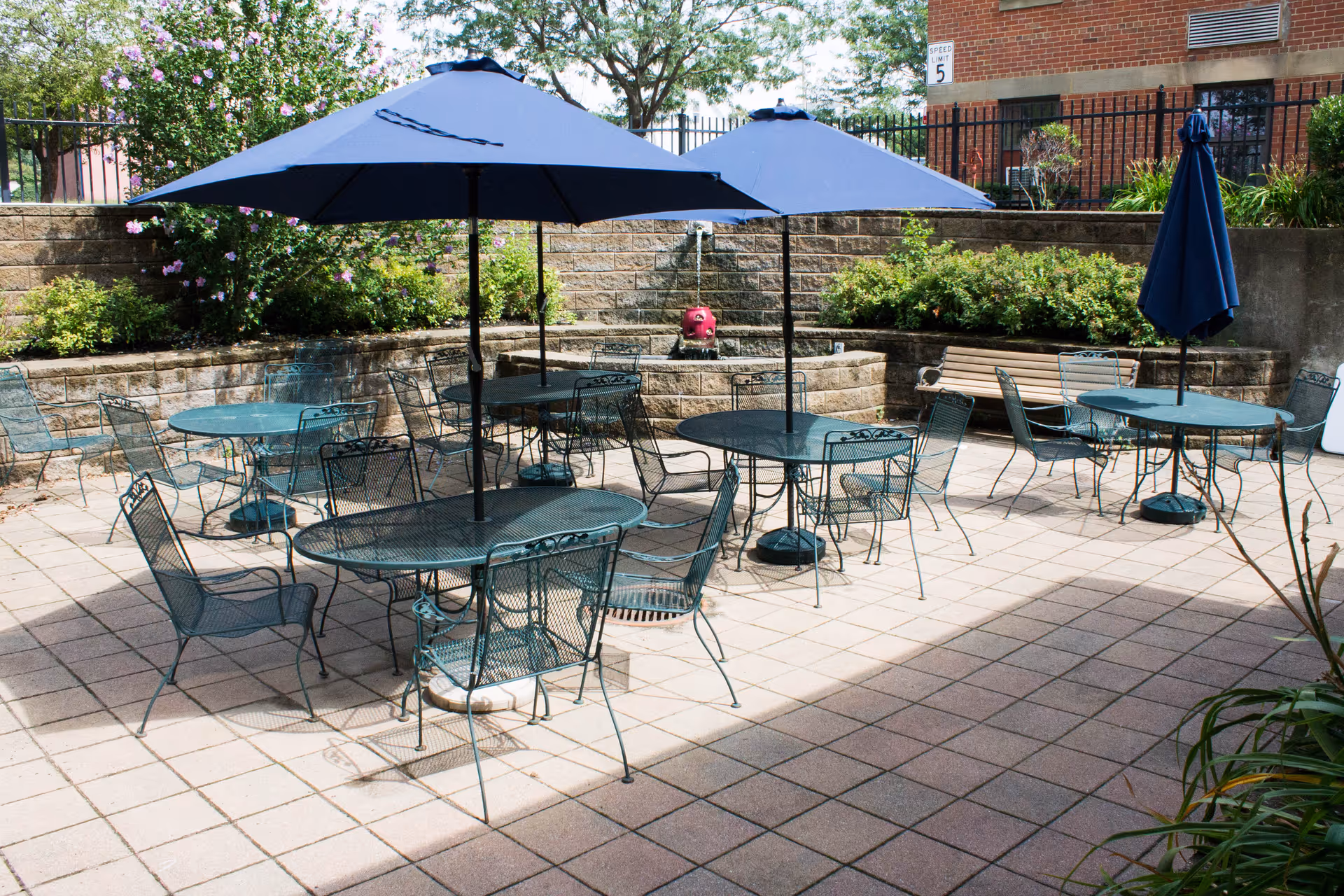 Outdoor patio area with several metal tables and chairs, each table shaded by a large blue umbrella. The patio is surrounded by a low stone wall with green bushes and flowers, and a brick building is visible in the background.