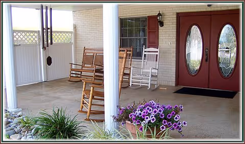 Covered porch area with two wooden rocking chairs and one white rocking chair, a hanging wind chime, potted purple flowers, and a double front door with oval glass panels.