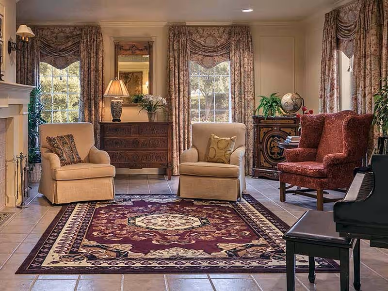 A cozy living room with two beige armchairs and one red armchair arranged around a patterned area rug. The room features large windows with floral curtains, a wooden chest with a lamp and flowers, a decorative cabinet with a globe and plants, and a piano in the foreground.